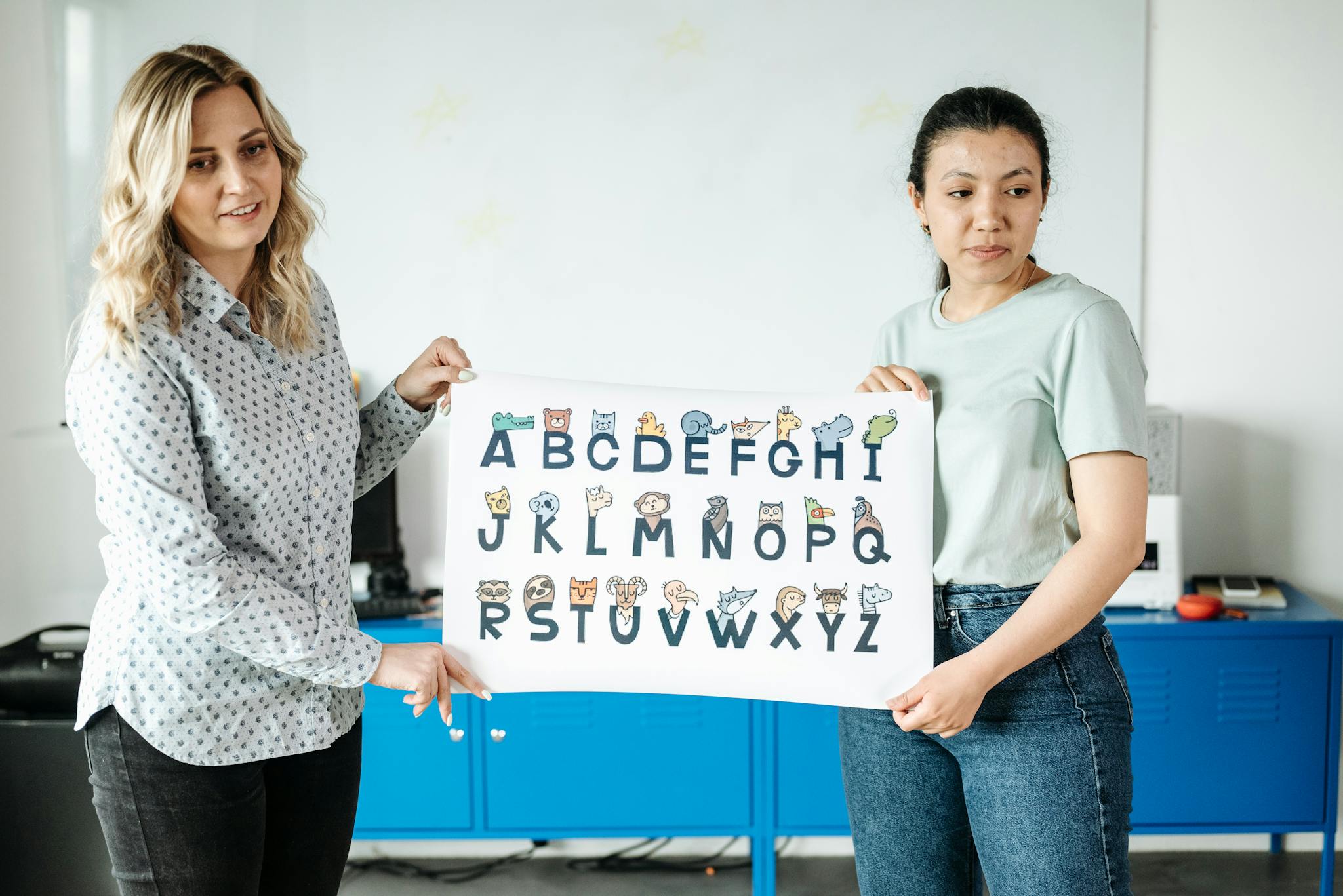 Two teachers hold an illustrated alphabet poster in a classroom, emphasizing education and learning.